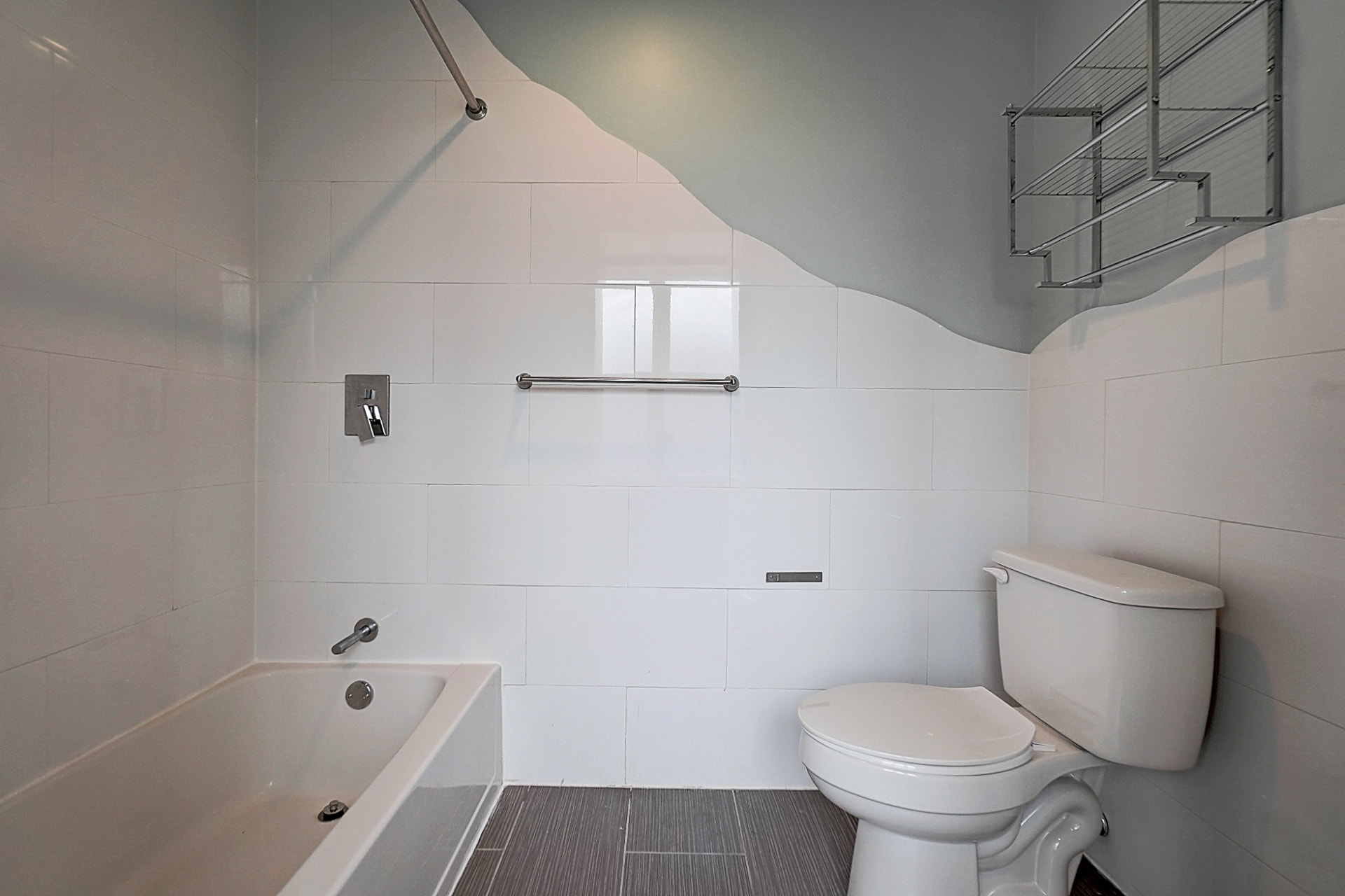 Bathroom with a bathtub on the left, a toilet on the right, white wall tiles, gray floor tiles and metal storage shelves.