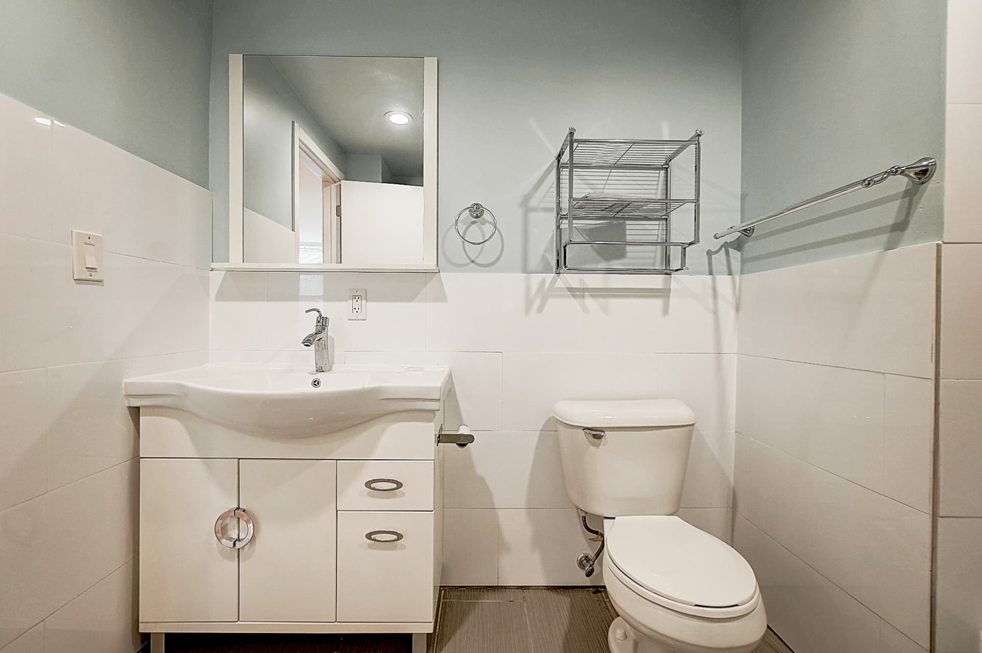 Bathroom with a white vanity and mirrored cabinet beside a toilet, metal wall shelves and a towel rail above white wall tiles.