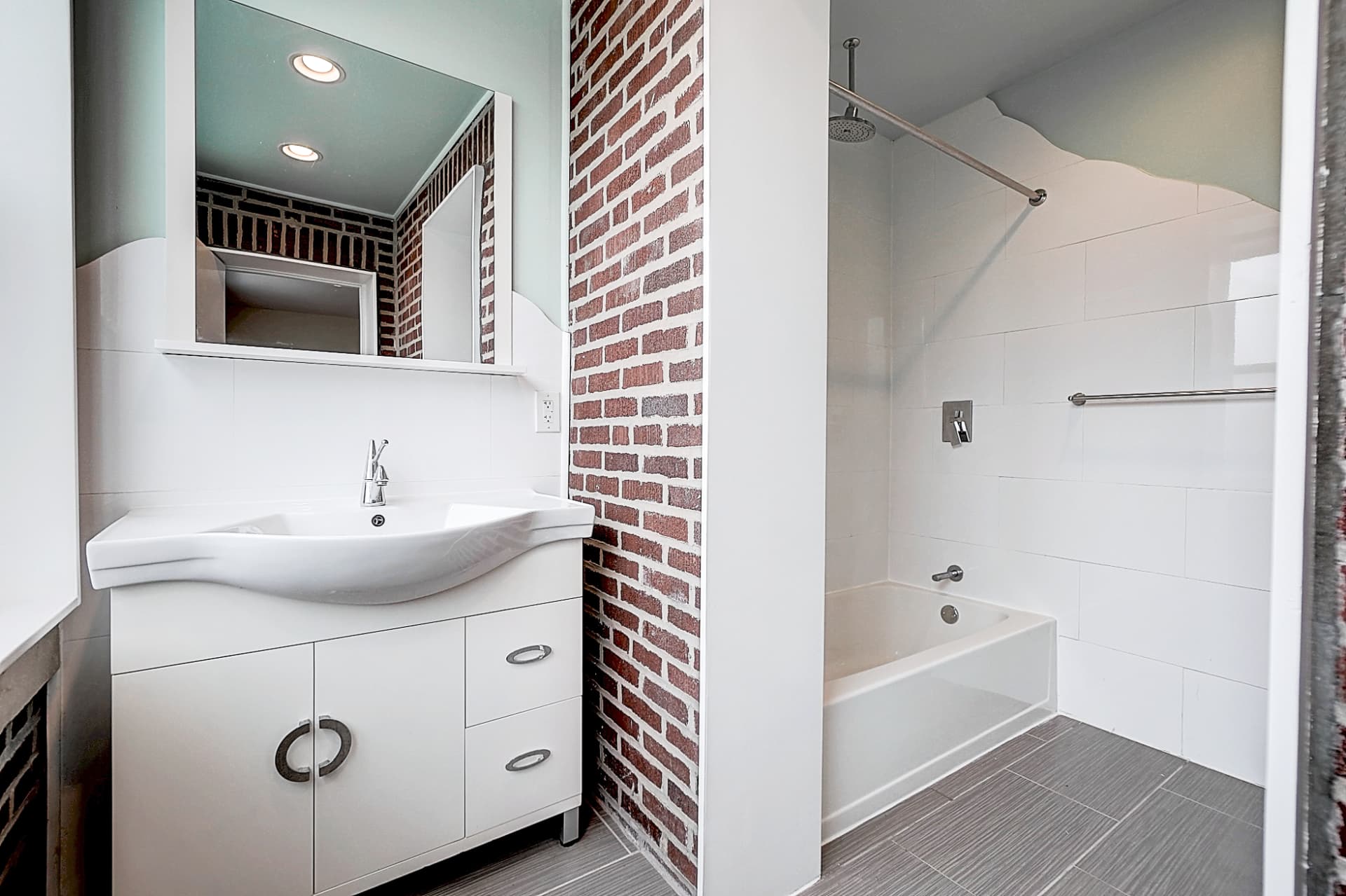 White bathroom vanity beneath a mirrored cabinet, beside an exposed brick wall and a bathtub with white tile surrounds.