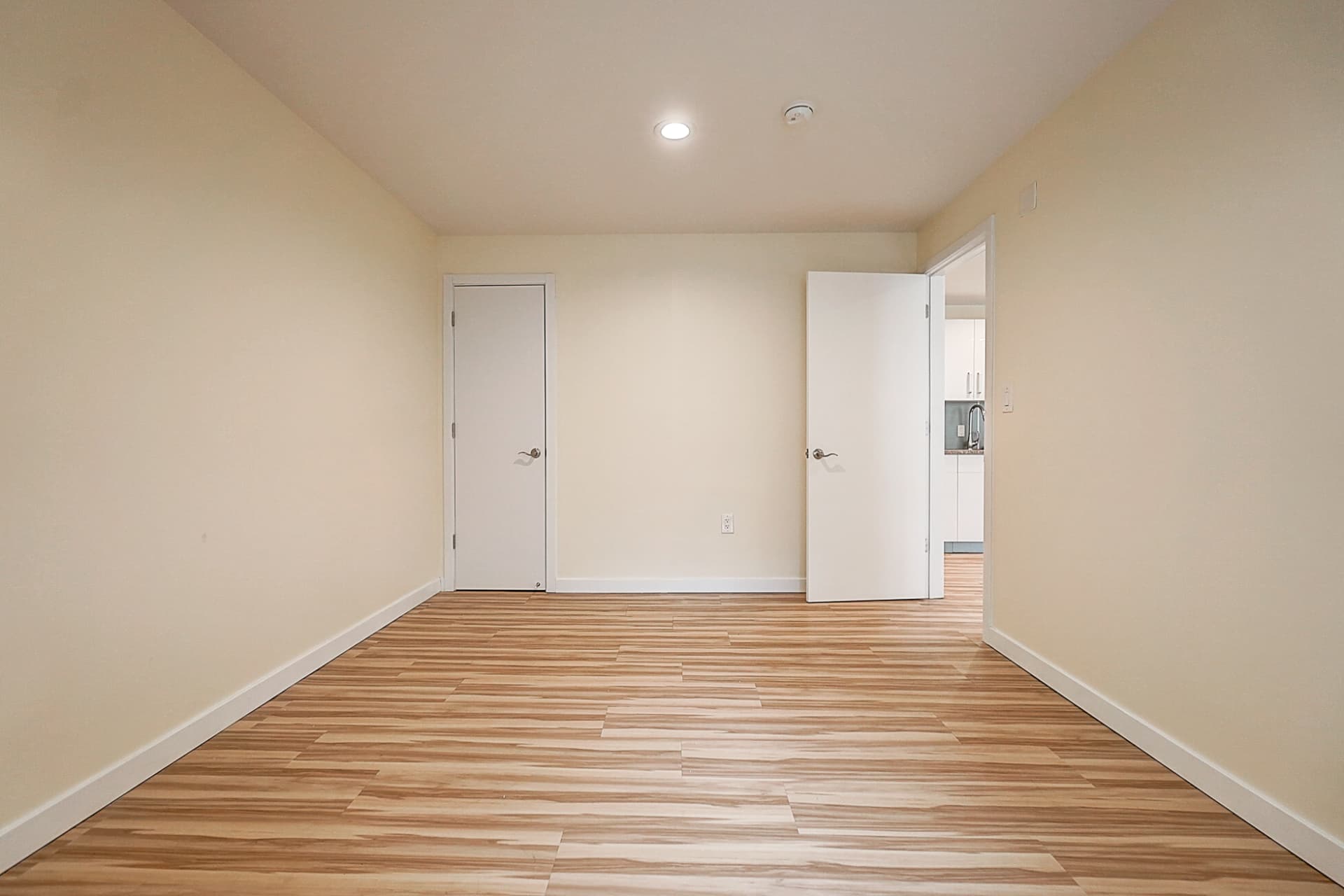 Long empty room with wood-pattern flooring, a closed door at the far end and an open doorway offering a view of white kitchen cabinets.