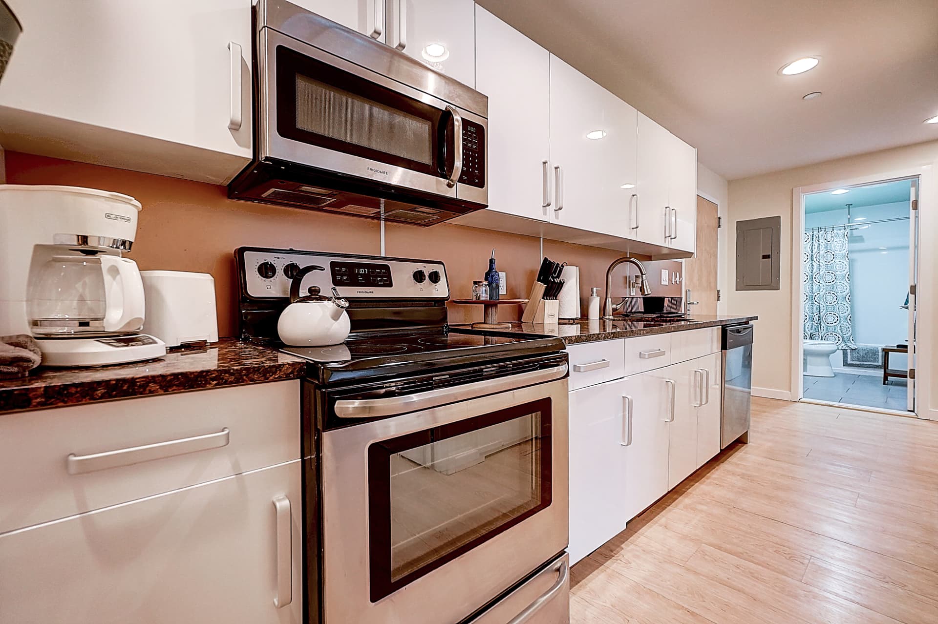 Angled view along a furnished kitchen with white cabinets, a stainless steel stove and microwave, countertop coffee maker and a bathroom doorway beyond.