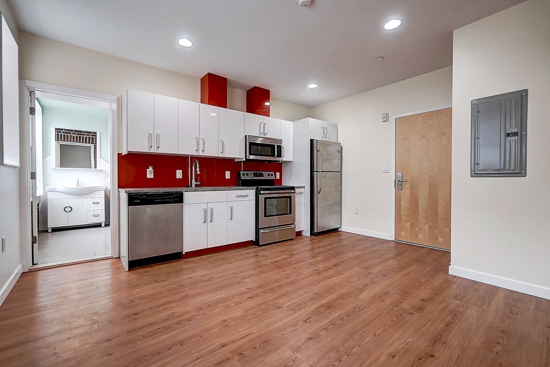 Open kitchen with white cabinets, a red backsplash, stainless steel appliances and wood flooring; a bathroom doorway is on the left.