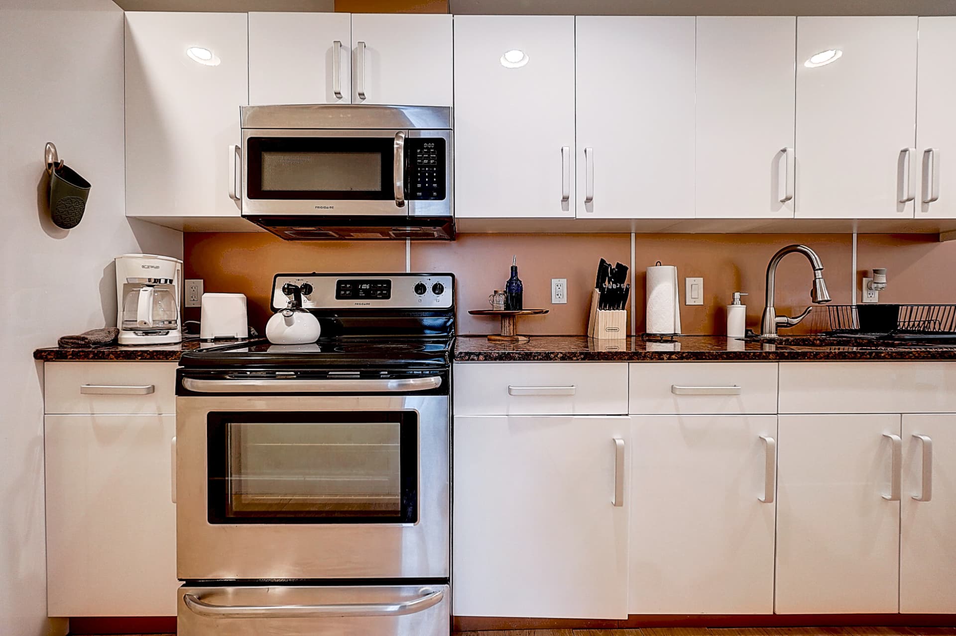 Front view of a kitchen with white cabinets, a stainless steel stove and microwave, dark countertop, coffee maker, kettle and sink.